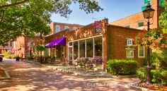 an old brick building with tables and chairs on the sidewalk in front of it is surrounded by greenery