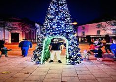 a man standing in front of a giant christmas tree with lights on it's sides