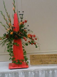 a tall red vase with flowers and greenery on top of a white table cloth