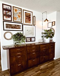 a wooden dresser sitting next to a wall filled with pictures and potted greenery