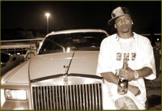 a man standing in front of a car holding a bottle and wearing a baseball cap