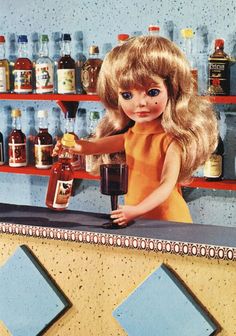 the doll is holding a blender in one hand and standing behind a counter with bottles on it