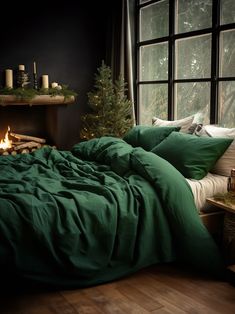 a bed with green sheets and pillows in front of a window next to a lit christmas tree