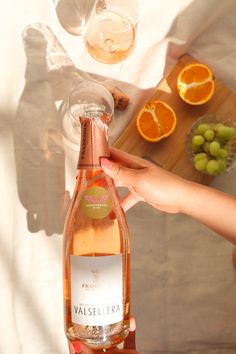 a person holding a bottle of wine in front of some oranges and grapes on a table