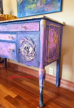 an old dresser painted purple and gold with flowers on the door knobs is sitting in front of a painting