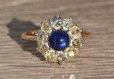 The Tana Antique Mixed Metal Sapphire Cabochon and Old Mine Cut Diamond Halo Ring. Crafted in 14 karat yellow gold and silver, this ring centers on a 1.50 carat royal blue cabochon sapphire. Surrounding the center cabochon sapphire is 1 carat total weight of chunky old mine cut diamonds in a round halo.  The diamonds have a clarity grade of VS2-I1 and a color grade ranging from O-P-Fancy Yellow.  This ring is a ladies finger size 7.5 and is able to be resized to any finger size. Each piece has b Yellow Gold Diamond Cabochons Fine Jewelry, Yellow Gold Multi-stone Cabochons For Fine Jewelry, Yellow Gold Multi-stone Cabochons Fine Jewelry, Vintage Yellow Gold Gemstone Cabochons, Elegant Yellow Gold Cabochons For Wedding, Vintage Yellow Gold Cabochons As Gift, Vintage Yellow Gold Cabochons For Gift, Vintage Gold Sapphire Ring With Cabochon, Vintage Gold Sapphire Cabochon Ring