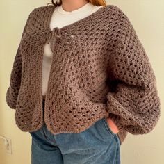 a woman wearing a brown crochet sweater and jeans standing in front of a wall