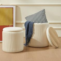 two white stools sitting on top of a wooden floor next to a painting and a black and white checkered pillow