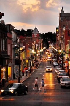 a city street with cars and people walking on the sidewalk at night time, as the sun sets