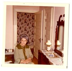 an older woman sitting in front of a mirror next to a dresser and table with flowers on it