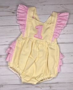 With ruffled shoulders and ruffles on the bottom, this is the most darling little sunsuit ever! Made of a pretty yellow seersucker with pink seersucker ruffles, it is cool and sweet for spring and summer. Available with numbers 1-5. Please indicate the number in the notes to seller section at checkout. Choose from ruffled shoulder straps that tie in the back with a bow, or that button at the waistband. Shoulders straps can have a single layer of ruffles, or a double layer of ruffles. The back ca Spring Cotton Bubble Romper With Ruffle Sleeves, Spring Cute Bubble Romper With Ruffle Sleeves, Summer Bubble Romper With Ruffles For Beach, Sweet Fitted Bubble Romper For Spring, Cotton Bubble Romper With Ruffle Sleeves For Summer, Summer Cotton Bubble Romper With Ruffle Sleeves, Pink Bubble Romper For Summer Birthday, Fitted Fun Bubble Romper For Spring, Spring Fitted Fun Bubble Romper