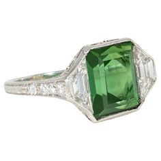 An Art Deco tourmaline, diamond, and platinum ring featuring an emerald-cut green tourmaline center flanked by trapezoid diamonds, with diamond-set shoulders and open scrolled foliate work in the gallery, in platinum. Marcus & Co. Atw tourmaline 4.61 cts., atw diamonds 1.75 cts. * Includes letter of authenticity for insurance purposes * Free shipping * Please do not hesitate to request additional photos of specific details or of this gorgeous period ring on a live model. Green Platinum Gemstones Fine Jewelry, Fine Green Platinum Gemstones Jewelry, Green Three Stone Platinum Ring, Gia Certified Green Emerald Art Deco Ring, Art Deco Green Ring, Gia Certified, Art Deco Green Ring Gia Certified, Art Deco Green Emerald Cut Diamond Ring, Green Emerald-cut Art Deco Diamond Ring, Fine Jewelry Green Diamond Platinum Ring