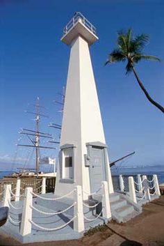 a tall white tower with a boat in the water behind it and a palm tree next to it