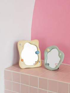 two small mirrors sitting on top of a pink tiled counter next to each other in front of a pink wall