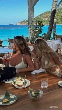 two beautiful women sitting at a table with food and drinks in front of the ocean