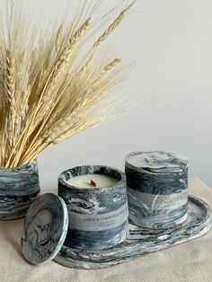 two candles sitting next to each other on top of a cloth covered tray with wheat stalks in the background