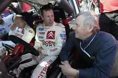 two men sitting in the drivers seat of a race car talking to each other while others look on