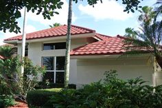 a white house with red tile roofing surrounded by trees and bushes in front of it