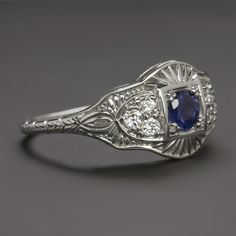 This beautiful and unique ring features a richl blue sapphire set in a diamond studded vintage style design that will never go out of style! The sapphire has a richly saturated blue hue that contrasts strikingly with the 14k white gold setting. Utterly classic and romantic, the antique style setting is richly textured with intricate filigree and milgrain. The effect is chic but also dreamy and softly feminine, a perfect choice for a timelessly stylish right hand ring or alternative engagement ri Classic Sapphire Halo Ring, Classic Sapphire Birthstone Ring, Classic Blue Sapphire Ring In 14k White Gold, Art Deco Blue Diamond Ring With Accents, Art Deco Blue Sapphire Ring With Center Stone, Luxury Blue Diamond Ring With Single Cut Diamonds, Art Deco Blue Diamond Ring With Center Stone, Classic Royal Blue Round Sapphire Ring, Blue Art Deco Diamond Ring With Center Stone