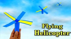 a hand holding a blue and yellow paper airplane in the sky with two small planes flying overhead