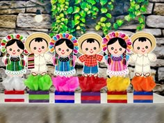 paper dolls are lined up on top of colorful tissue pom poms in front of a stone wall