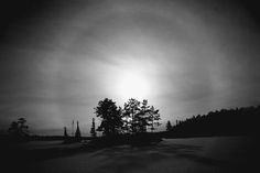 black and white photograph of sun shining through the clouds over trees in an open field