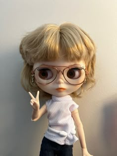 a doll with blonde hair and glasses giving the peace sign while standing next to a wall