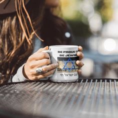 a woman sitting at a table with a coffee mug in her hand and the words be proud be jewish on it