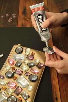 two hands holding a tube of glue over a wooden table with various jewels on it