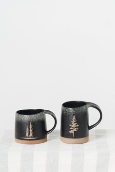 two black mugs with trees on them sitting on a tablecloth covered table cloth