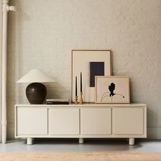 a white cabinet with two pictures on it and a lamp next to it in front of a brick wall