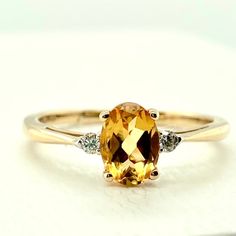 We're Obsessed With The Beautiful Color Of This Citrine Stone! Made Of 14kt Yellow Gold This Ring Has A Standard Straight Yellow Gold Band With Slightly Curved Edges That Sits Comfortably On Your Finger. In A Slightly Raised Setting Sits A 5mm X 7mm Oval Cut Yellow And Orange Toned Citrine Stone Held Securely By Four Prongs. On Either Side Of This Center Stone Sits A Diamond; These Diamonds Come To A Total Carat Weight Of 0.05. This Make Them Nicely Sized Accent Stones That Allow The Citrine To Be The Center Focus. This Ring Is Incredibly Comfortable And Durable Enough For Regular Every Day Wear. Classic Citrine Ring With Center Stone, Citrine Ring With Center Stone In Round Cut, Citrine Jewelry With Accent Stones In Round Cut, Fine Jewelry Citrine Ring With Round Cut, Gold Citrine Diamond-cut Jewelry, Citrine Jewelry, Citrine Stone, Gold Band Ring, Oval Cut