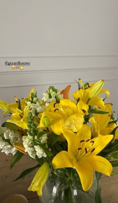 a glass vase filled with yellow flowers on top of a wooden table in front of a white wall