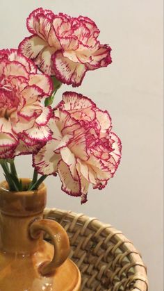 three pink flowers in a brown vase on a wicker tablecloth with a white wall behind it