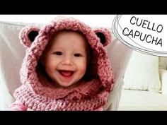 a smiling baby wearing a pink knitted bear hat and scarf with the caption cueello capucha