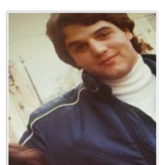 a man in a blue jacket and white turtle neck sweater posing for the camera with his hands on his hips