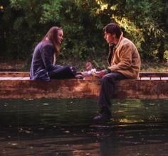 two people sitting on the edge of a body of water with trees in the background