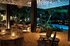 people are sitting at tables on the sand in front of a swimming pool and palm trees