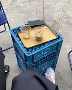 two cups of coffee sitting on top of a blue crate next to a person's legs