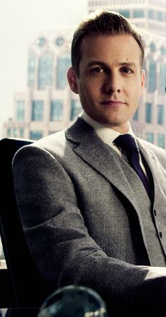 a man wearing a suit and tie sitting in front of a window with a cityscape behind him