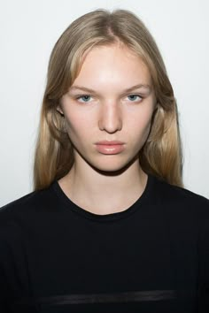 a woman with long blonde hair standing in front of a white wall and looking at the camera