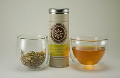 a cup of tea next to a canister and glass with liquid in it on a white surface