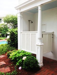 an outdoor shower in the corner of a white house with brick walkway and green plants