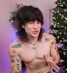 a man with long hair and tattoos on his chest is talking to someone in front of a christmas tree