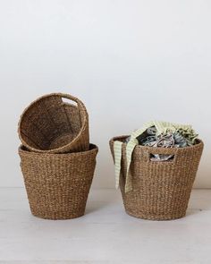 A still life image features two Round Woven Baskets by Creative Co-op placed against a plain white background. The basket on the left is empty with another basket nested inside it, showcasing their natural charm. The basket on the right contains rumpled, colorful fabric with green and white striped material on top, emphasizing an eco-friendly aesthetic. Road Trip Gifts, Seagrass Baskets, Grass Basket, Towel Organization, Seagrass Basket, Hand Woven Baskets, Basket Set, Home Goods Decor, Creative Co Op