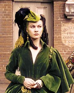 a woman in a green dress and feathered headdress is standing by a brick building