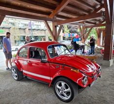 an old red car is parked under a wooden structure and people are standing around it