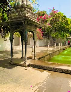 a gazebo in the middle of a park with water running through it and flowers on the ground