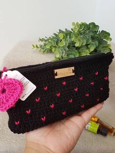 a hand holding a black crocheted purse with pink hearts on it and a bottle of essentials next to it