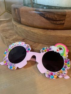 two pink sunglasses with rainbow accents on them sitting on a wooden table next to a potted plant